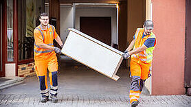 2 Männer in Arbeitskleidung tragen ein Möbelstück aus einem Gebäude.