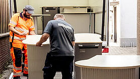 Zwei Mitarbeiter des Gebrauchtwarenkaufhauses, einer in orangener und einer in dunkler Arbeitskleidung beugen sich über ein Möbelstück